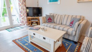 A living room with a striped sofa, a white coffee table, a flat-screen TV on a wooden stand, and a large window with curtains. - Südstrand 3, Germany (Sylt)