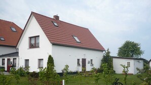 Une maison blanche avec un toit rouge, une petite cour avec un banc et un jeune arbre.