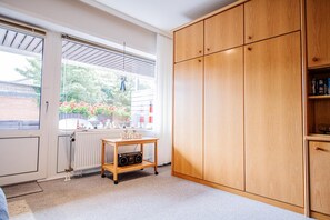 Una habitación con un armario de madera, un radiador, una ventana con vistas a un jardín y una pequeña mesa de madera con un radio.