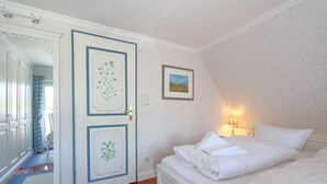 Et soveværelse med en seng, en dør med blomstermotiver, et billede i ramme på væggen og et natbord med en lampe.