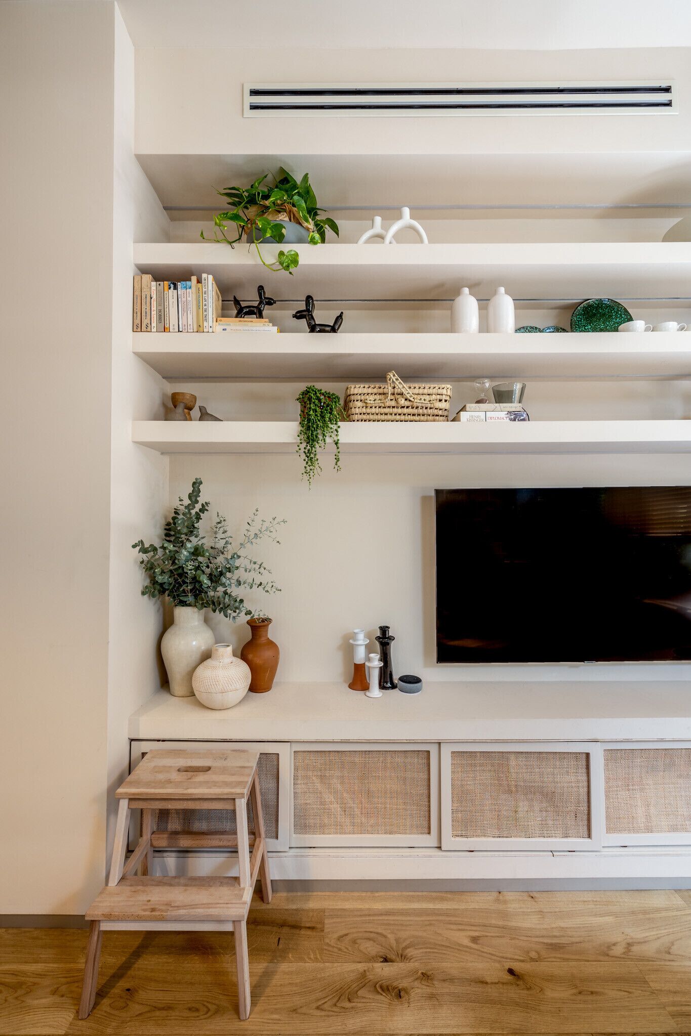 A modern living room with a mounted TV, white shelves with decorative items, and a wooden floor.