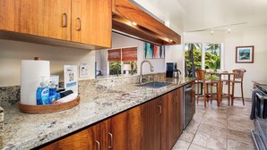 A kitchen with wooden cabinets, a granite countertop, a coffee maker, and a dining area with a table and chairs. - Kanaloa at Kona 1606: Beautiful Oceanview Condo w/ Shared Pool, Spa, AC & Lanai (Kailua Kona)