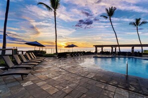 A poolside area with lounge chairs and umbrellas, overlooking a sunset over the ocean.