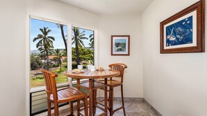 A room with a round wooden table and chairs, a window with a view of palm trees and a distant landscape, and two framed pictures on the wall. - Kanaloa at Kona 1606: Beautiful Oceanview Condo w/ Shared Pool, Spa, AC & Lanai (Kailua Kona)