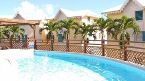 A swimming pool with a wooden deck and palm trees, surrounded by buildings with a white and yellow color scheme. - Charming apartment with swimming pool in secure residence and water tank (Sainte-Anne)
