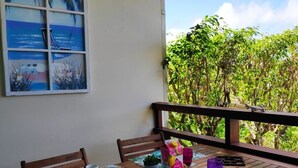 A wooden dining table with chairs, a tropical-themed mat, and a view of lush greenery through a window. - Charming apartment with swimming pool in secure residence and water tank (Sainte-Anne)