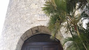 A stone building with a large wooden double door, a sign above the door, and a palm tree in the foreground. - Charming apartment with swimming pool in secure residence and water tank (Sainte-Anne)
