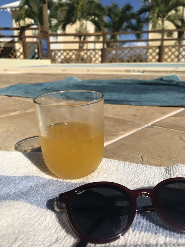 A glass of yellowish beverage on a towel by a poolside, with sunglasses and a blue towel nearby. - Charming apartment with swimming pool in secure residence and water tank (Sainte-Anne)