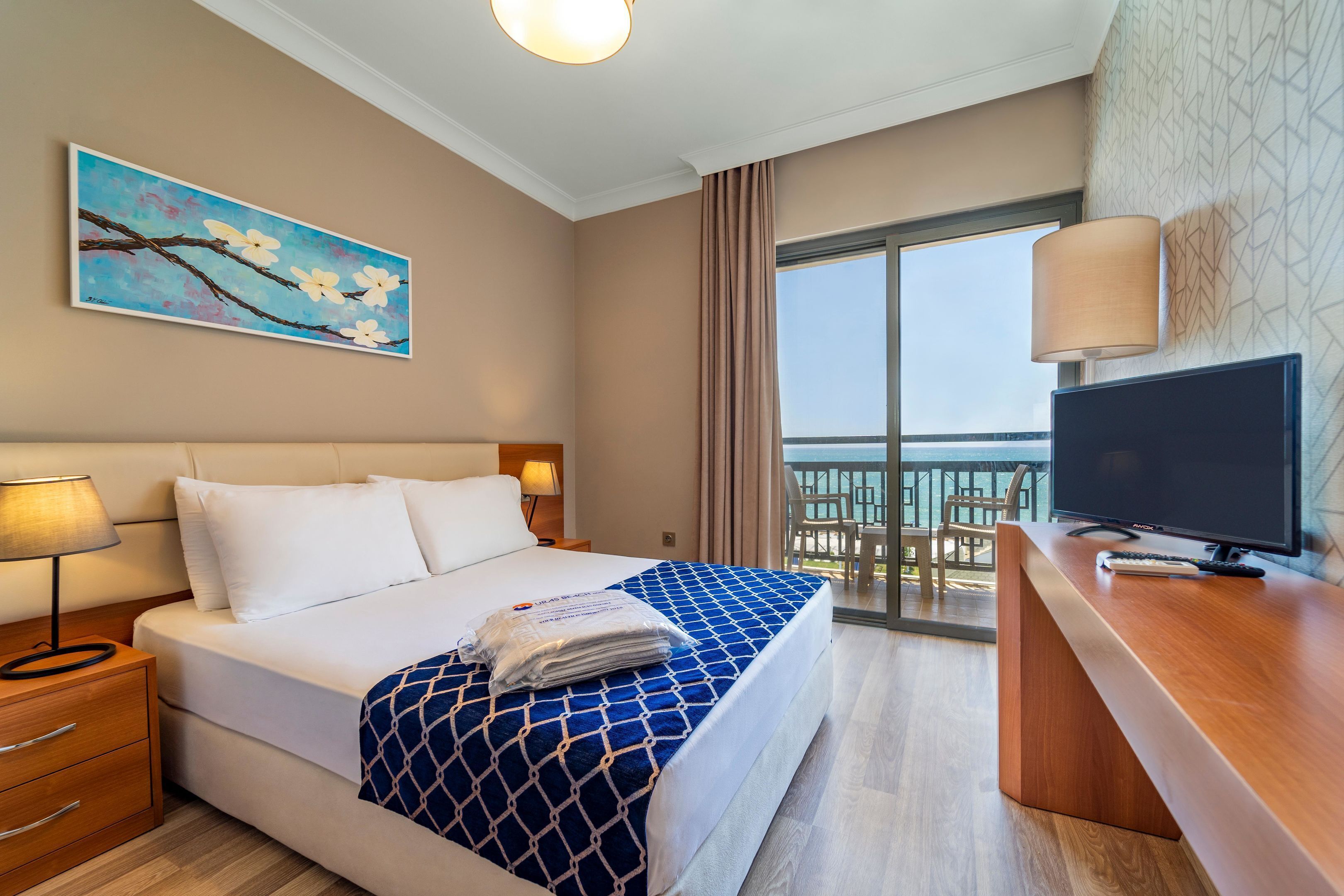Habitación de hotel con cama, escritorio con computadora, televisor, un cuadro en la pared y balcón con vista.