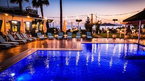 A swimming pool with lounge chairs and string lights, surrounded by a landscaped area with palm trees and a view of the sea.