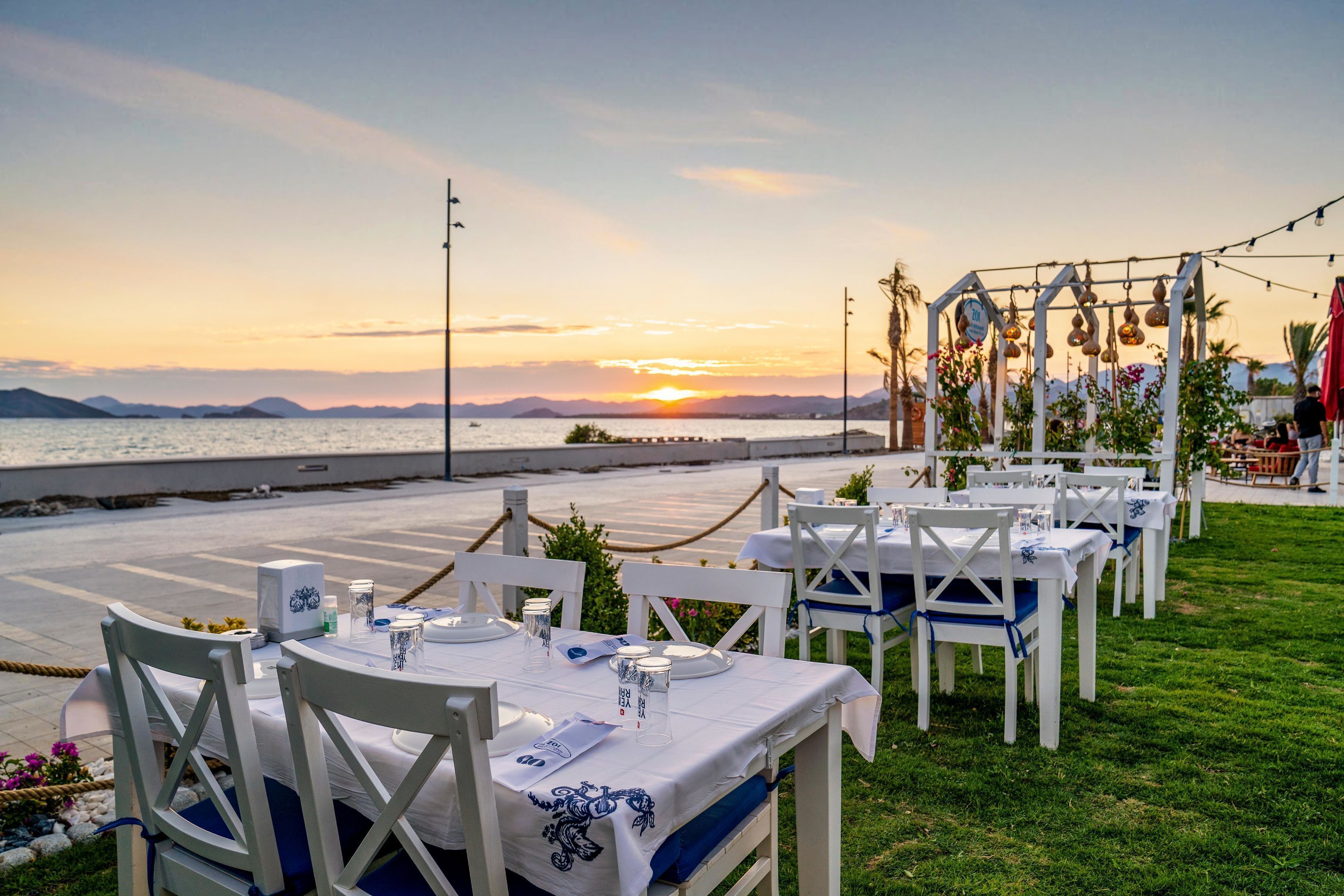An outdoor dining area with white tables and chairs, set against a backdrop of a sunset over the sea.