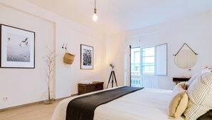 Quarto com cama grande, piso de madeira, quadro com passarinhos, bolsa pendurada, tripé e espelho redondo na parede.