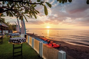 A beachside setting with tables and chairs, a row of kayaks, and a sunset over the ocean.