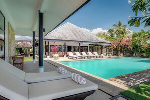 A poolside area with lounge chairs, a covered seating area, and a view of a building with a tiled roof.