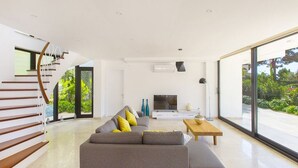 A modern living room with a grey sofa, wooden staircase, and a large window offering a view of greenery.