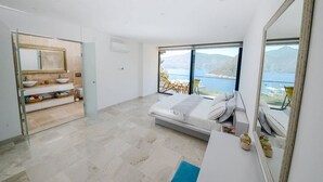 A bedroom with a large bed, a bathroom with a vanity, and a view of the sea.