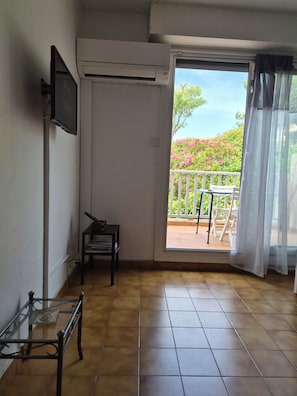 A room with a tiled floor, a sliding glass door leading to an outdoor balcony, a mounted television, and a small table with books. - Comfortable, serene studio in residence, swimming pool, tennis court, private parking, terrace. (Sausset-les-Pins)
