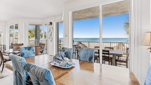 A spacious dining area with a wooden table and chairs, large windows offering a view of the ocean, and a balcony with a view of the beach.