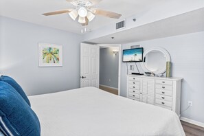Ein Schlafzimmer mit Bett, Deckenventilator, Kommode, Fernseher und einer Tür zu einem anderen Raum.