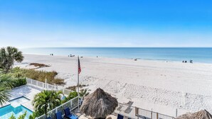 A beach view with a swimming pool, a thatched umbrella, and a flag. - Sand Isle II C—Beachfront Beauty w/ Pool, Sunset Balcony & Walkable Dining (Indian Rocks Beach)
