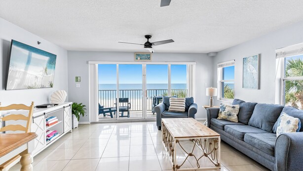 A spacious living room with a large window, a ceiling fan, a wooden coffee table, and a grey sofa. - Sand Isle II C—Beachfront Beauty w/ Pool, Sunset Balcony & Walkable Dining (Indian Rocks Beach)