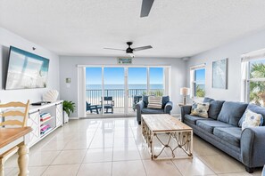 A spacious living room with a large window, a ceiling fan, a wooden coffee table, and a grey sofa. - Sand Isle II C—Beachfront Beauty w/ Pool, Sunset Balcony & Walkable Dining (Indian Rocks Beach)