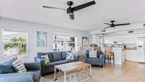 A spacious living room with a sofa, a coffee table, and a dining area with chairs. There are ceiling fans and a kitchen in the background. - Sand Isle II C—Beachfront Beauty w/ Pool, Sunset Balcony & Walkable Dining (Indian Rocks Beach)