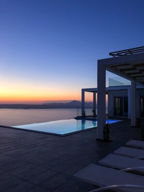 Un espace piscine extérieur moderne offrant une vue dégagée sur la mer et les montagnes lointaines, particulièrement au coucher du soleil.