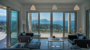 Una sala de estar moderna con un gran ventanal que da a un balcón, con vistas panorámicas al mar y a las montañas.