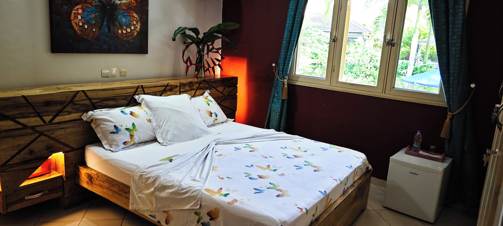 A bed with white bedding, a bedside table with a lamp, a plant, and a window with curtains.
