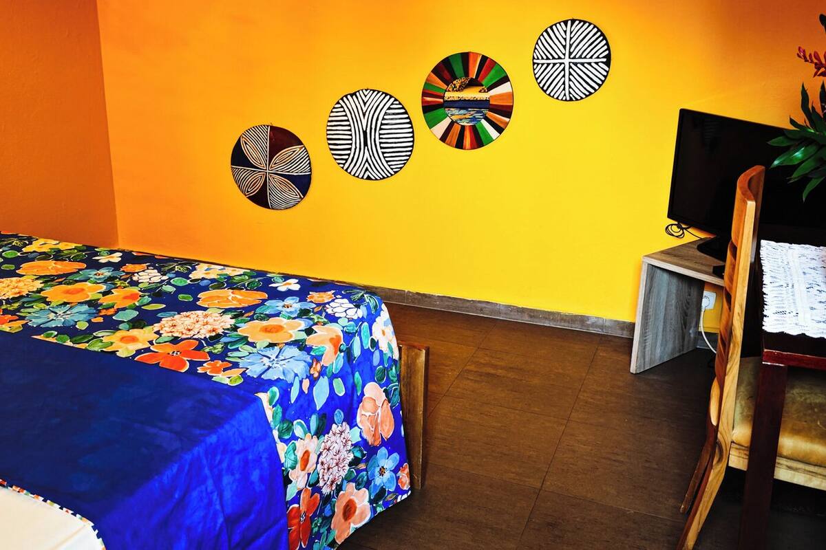 A room with a bed, a desk, a chair, a plant, and wall decorations.