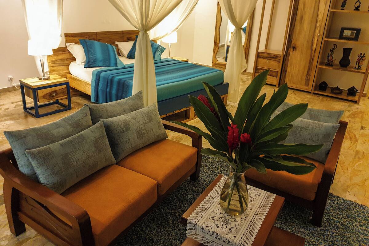 A bedroom with a bed, a wooden nightstand, a bedside table, a sofa, and a decorative plant.