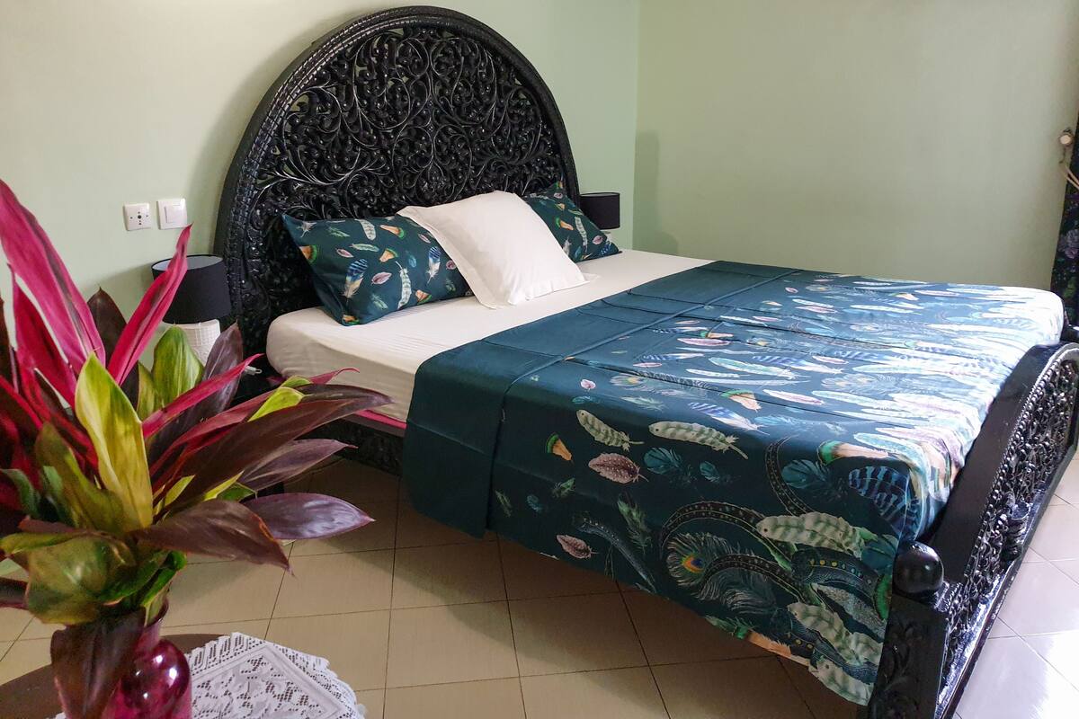 A bedroom with a bed, a nightstand, a lamp, a potted plant, and an air conditioning unit.
