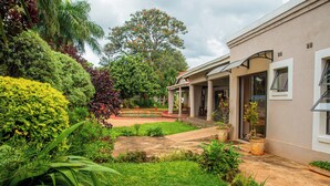 A house with a covered porch, a garden with various plants, and a large tree in the background. - The Berkshire (Harare)