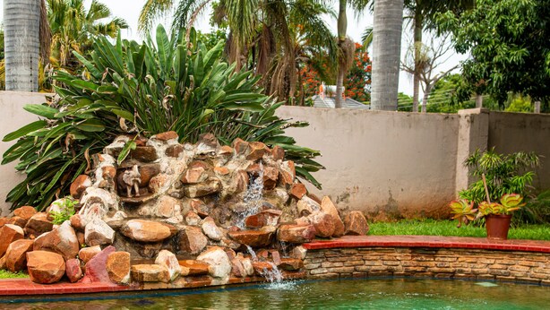 A pool with a rocky waterfall feature surrounded by tropical plants and a concrete wall. - The Berkshire (Harare)