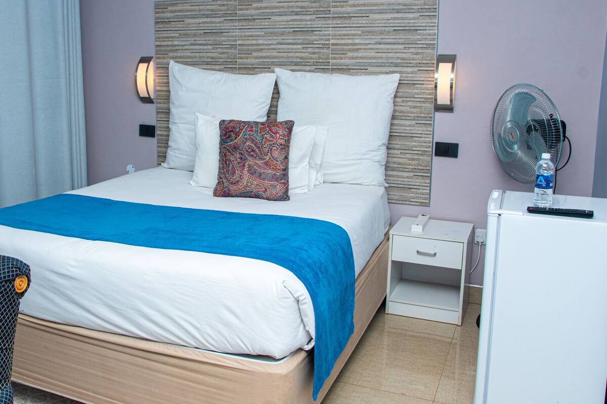 Un lit bien fait, avec du linge de lit blanc et un plaid bleu, une table de chevet avec une télécommande, et un ventilateur fixé au mur.