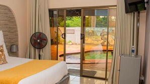 A hotel room with a bed, a fan, a water bottle, and a sliding glass door leading to an outdoor area with a pool and greenery. - The Berkshire (Harare)