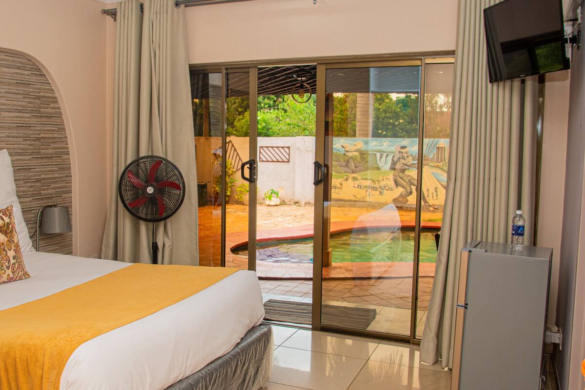 Une chambre d’hôtel équipée d’un lit, d’un ventilateur, d’une bouteille d’eau et d’une porte-fenêtre coulissante donnant sur un espace extérieur avec une piscine et de la verdure.