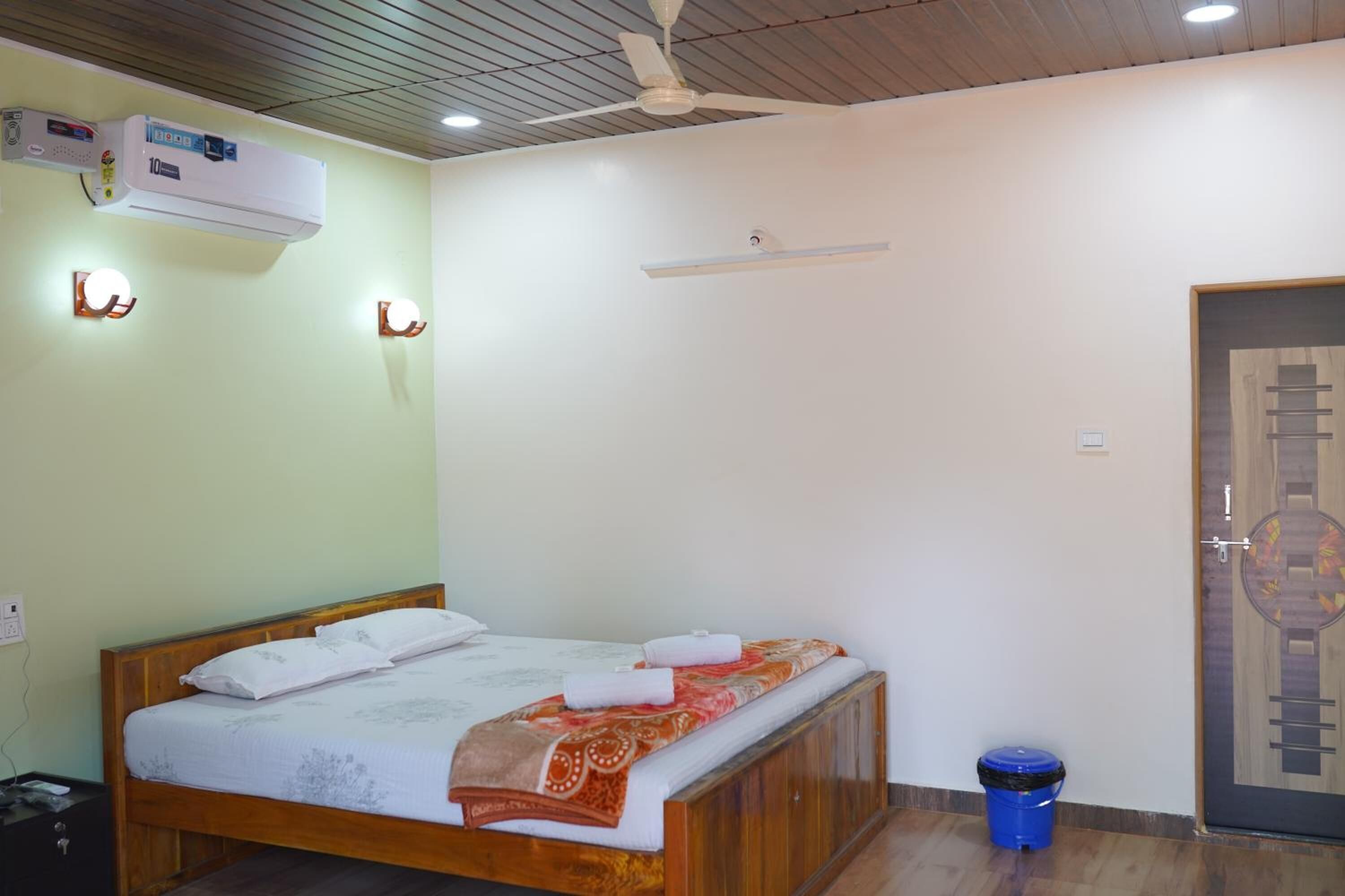 Una cama individual con estructura de madera, ropa de cama blanca y una colcha estampada. Se observa un ventilador de techo, un equipo de aire acondicionado y un cubo de basura azul.
