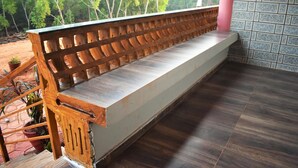 A wooden bench with a slatted backrest and a flat seat, placed on a tiled floor with a view of trees through a window. - Viraz Valley (Kumta)