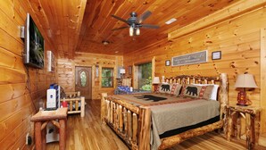 A wooden cabin bedroom with a bed, a ceiling fan, a TV, and a small table with a lamp.