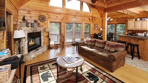 A cozy log cabin interior with a stone fireplace, wooden furniture, and a large window overlooking the outdoors.