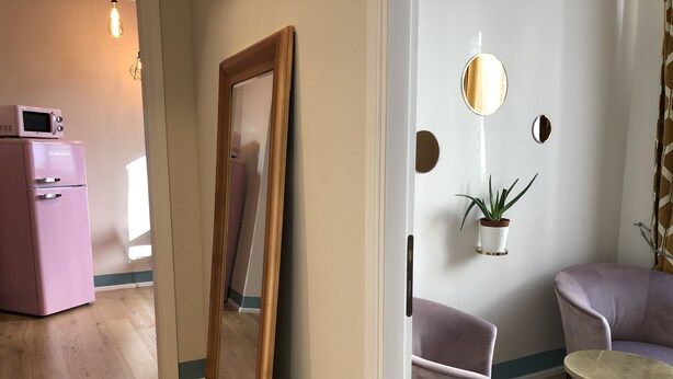Ein modernes Wohnzimmer mit Holzboden, einem pinken Kühlschrank, einem großen Spiegel, einem violetten Sessel, einem runden Tisch und einer Wandpflanze.
