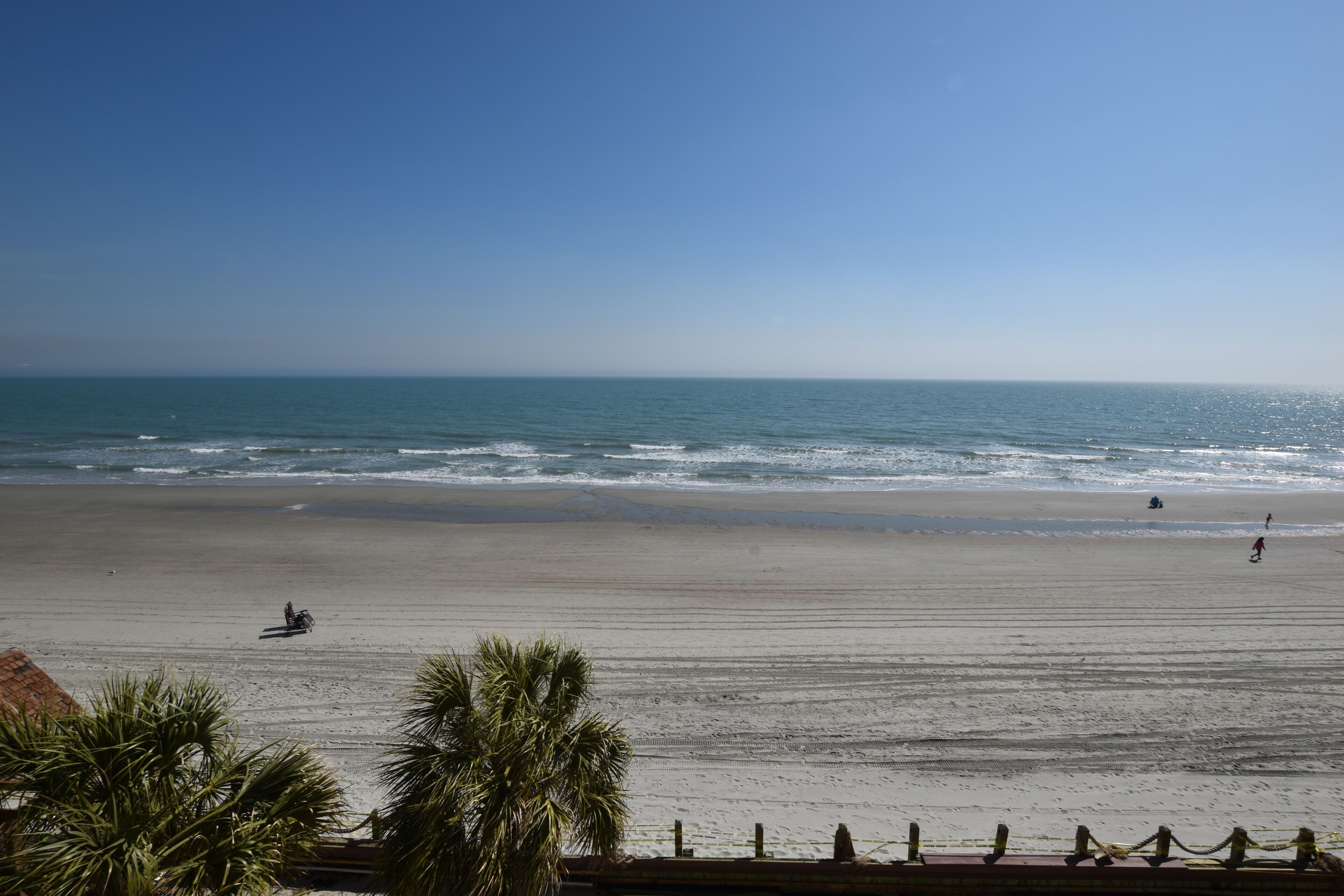 En strand med klar himmel, stilla hav och några få personer som promenerar.