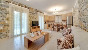 A modern living room with a sofa, coffee table, and a TV. There is a kitchen area with a dining table and chairs. - Villa Nikos - Faraggi Villas (Zoniana)