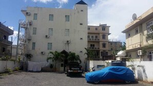 A multi-story building with a flat facade, a satellite dish, and a covered vehicle in the foreground. - Hôtel le corail (Port Louis)
