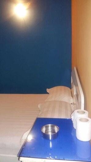 A small room with a bed, a blue table with a metal bowl and two rolls of toilet paper. - Hôtel le corail (Port Louis)