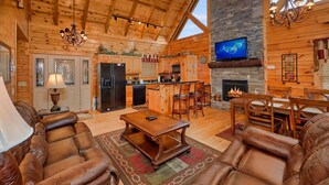 Interior de una cabaña de troncos con sala de estar, comedor, cocina, chimenea y televisor montado en la pared.