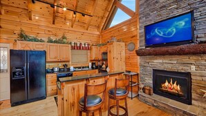 Una cocina al estilo de cabaña de troncos con chimenea de piedra, taburetes de madera y un televisor colocado sobre la chimenea.