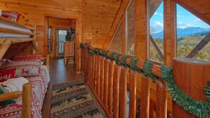 A log cabin interior with a wooden staircase, a bed with a red and white patterned blanket, and a view of mountains through large windows. - Reindeer Bay: Private Pool, Amazing Views, Games (PIgeon Forge)